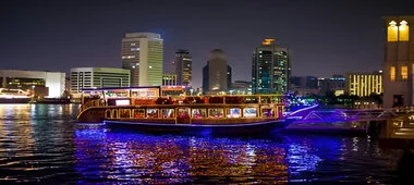 An illuminated cruise ship docked at a marina at night, with the city skyline reflecting on the water.